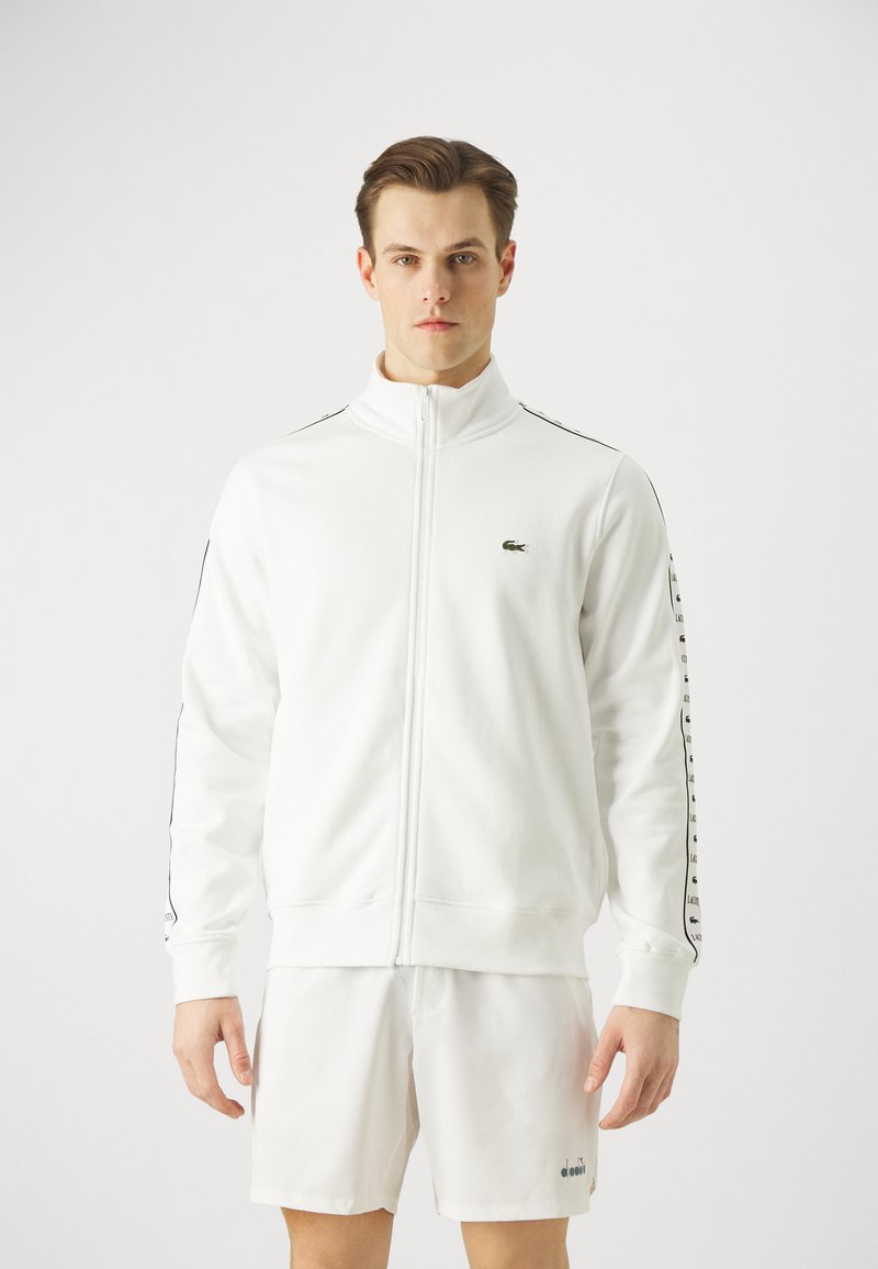 Witte ritsjack gemaakt van zachte stof, voorzien van een hoge kraag, zwarte zijstrepen en een groen logo op de borst. Draag het met bijpassende shorts.