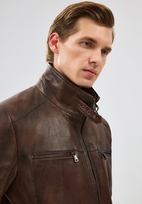 Veste en cuir marron avec un col haut, fermeture éclair à l'avant et deux poches latérales. Présente un matériau texturé et des accents de quincaillerie contrastants.