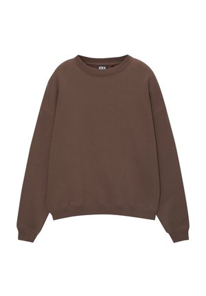 Person iført en brun oversized sweatshirt, lyseblå vaskede jeans og to perlehalskæder, med den ene hånd i lommen og den anden langs siden.
