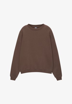 Personne portant un sweat-shirt marron oversize, un jean bleu clair délavé et deux colliers de perles, une main dans la poche et l'autre sur le côté.