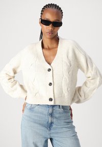 Cardigã em ponto trançado creme com decote em V, cinco botões pretos, comprimento ligeiramente cropped e punhos canelados, combinado com jeans azul claro.