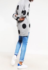 Šedý svetr s velkými černými puntíky, dlouhými rukávy a volnějším střihem. Nošený s světle modrými džínami a bílými teniskami s modrými akcenty.