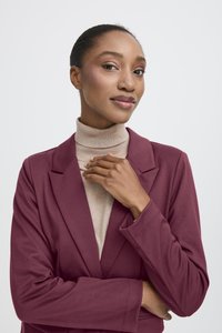 Mörkröd blazer med en skräddarsydd design, bärs över en beige polotröja. Slät textur med slagkrage och vikta manschetter.