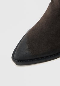 Barna nyersbőr bokacsizma hegyes orral és fekete bőr díszítéssel, texturált felülettel és varrott talprésszel.