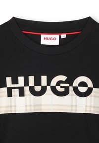 T-shirt noir arborant un logo "HUGO" en blanc, sur fond de tartan beige. Encolure ronde avec des détails de couture rouges.