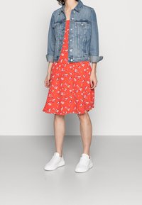 Robe fleurie rouge avec un ourlet évasé, assortie d'une veste en denim bleu clair. Des baskets blanches complètent la tenue. Textures lisses et style décontracté.