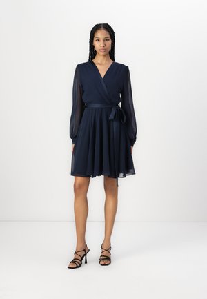 Navy wrap dress with long sheer sleeves, a tie waist, and a flared skirt. Features a deep V neckline and smooth texture. Black strappy heels.