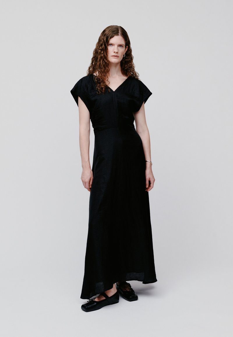 Γυναίκα με μακριά, σγουρά μαλλιά που φοράει μαύρο αμάνικο μάξι φόρεμα με V λαιμόκοψη και μαύρα πλατφόρμες, στέκεται μπροστά σε απλό φωτεινό φόντο.