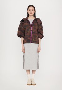 Gekürzte Tarnjacke mit einem rosa Reißverschluss und Klettverschluss-Akzenten, kombiniert mit einem grauen Midikleid mit Seitenschlitzen und beigen Sneakers.
