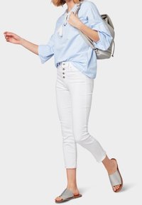 Himmelblaues Langarmoberteil mit Fransen-Detail, kombiniert mit weißen, knöchellangen Jeans mit Knopfakzenten und grauen Slip-On-Sandalen.