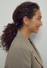 Curly hair in a ponytail secured with a gold accessory; wearing gold stud earrings. Dressed in a light brown blazer. Profile view.