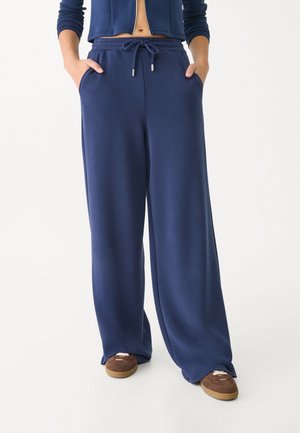 Personne portant un pantalon de survêtement bleu marine à jambes larges avec taille à cordon et baskets marron, debout les mains dans les poches.