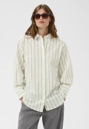 Cămașă albă cu dungi verzi verticale, design cu nasturi, mâneci lungi și tiv curbat. Purtați împreună cu pantaloni gri și ochelari de soare.