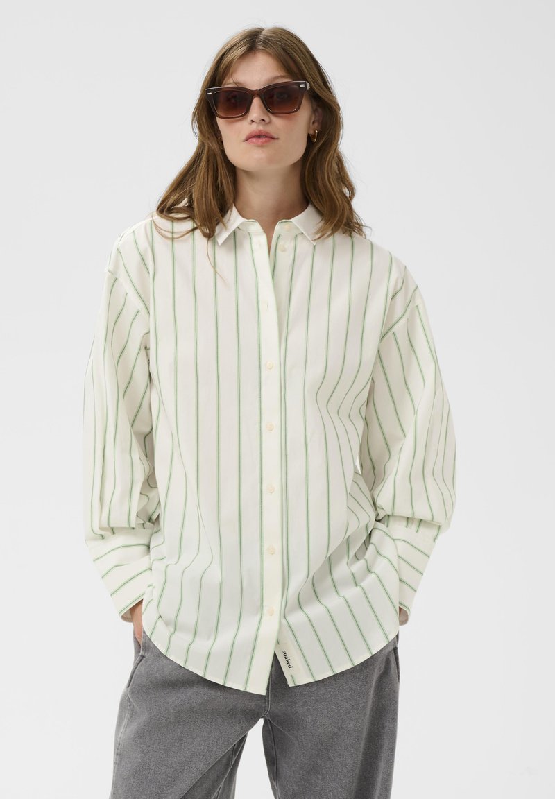 Chemise blanche à rayures avec des lignes verticales vertes, modèle boutonné, manches longues et ourlet arrondi. Portée avec un pantalon gris et des lunettes de soleil.