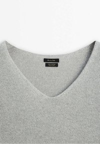 Pull gris clair en maille avec col en V et deux étiquettes en tissu noir au col, l'une indiquant le nom de la marque et l'autre le matériau et l'origine.