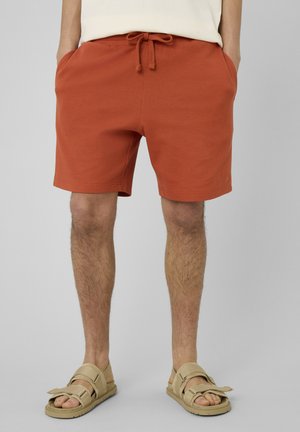 Homme portant un short à cordon couleur rouille avec des poches et des sandales beiges ouvertes, debout devant un fond gris clair uni.