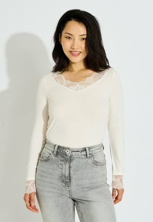 Mujer sonriente con cabello negro que lleva una camiseta blanca de manga larga con detalles de encaje y vaqueros de tiro alto de color gris claro sobre un fondo liso.