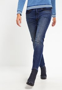 Tummansiniset farkut suoran leikkauksen ja pienen haalistuman kanssa. Yhdistetty vaaleansiniseen pitkähiharaiseen paitaan, jossa on valkoiset raidat hihansuissa.