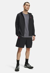Chaqueta ligera negra con capucha, camiseta gris y pantalones cortos negros con bolsillos tipo carga. Presenta acentos de logotipo en rojo y calzado deportivo.