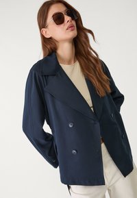 Námořnická modrá dvouřadá bunda s velkým klopovým límcem, se zapínáním na knoflíky a volným střihem, spárovaná s béžovým topem.