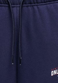 Marineblauwe trainingsbroek met elastische tailleband, trekkoordsluiting en een bedrukt logo in rood en wit. Katoenen stof met een gladde textuur.