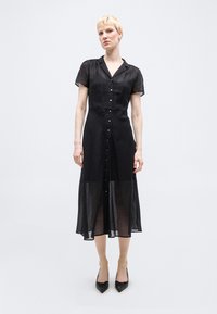 Γυναίκα με κοντά ξανθά μαλλιά, ντυμένη με μαύρο διάφανο midi φόρεμα με κουμπιά και μαύρες μυτερές γόβες, που στέκεται μπροστά σε απλό λευκό φόντο.