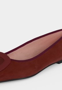 Gros plan sur une chaussure plate en daim bordeaux avec un bout arrondi, une boucle décorative et une doublure intérieure en tissu rose.