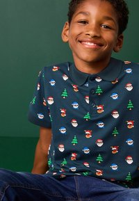Marinblå polo-shirt med ett lekfullt julmönster av tomteansikten, snögubbar och träd. Tyget verkar mjukt och har en klassisk krage.
