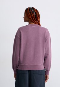 Woman with long orange braids wearing a loose mauve sweatshirt and dark pants, standing facing away against a plain light background.