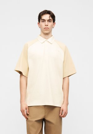 Fiatal, sötét hajú férfi, aki túlméretes, krémszínű és bézs rövid ujjú galléros inget és világosbarna nadrágot visel, sima háttér előtt áll.