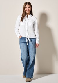Camicia bianca a maniche lunghe con bottoni, dotata di due tasche sul petto e vita legata, abbinata a jeans a gamba larga blu chiaro e scarpe beige.