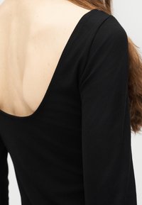 Γυναίκα με μακριά καστανά μαλλιά που φοράει μαύρο μακρυμάνικο τοπ με βαθύ ντεκολτέ στην πλάτη, σε απλό φόντο.