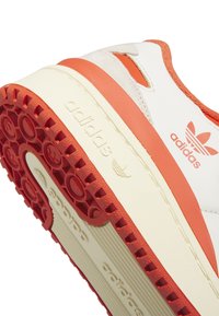 Adidas sporta apavi ar baltu augšdaļu un oranžiem akcentiem, struktūrizētu gumijas zoli un logotipa detaļu sānos un papēdī.