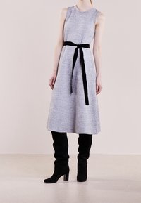 Robe grise sans manches avec une silhouette trapèze, ceinturée d'un ruban en velours noir. Portée avec des bottes noires en suède montant jusqu'aux genoux.