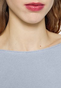 Pull en tricot bleu clair avec un large décolleté, une texture lisse et des coutures visibles ; les lèvres du modèle ont une teinte rose naturelle.