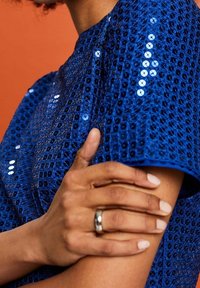 Personne portant un haut bleu à sequins avec une texture en filet, tenant leur bras supérieur, portant une bague en argent sur le majeur.
