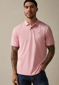 Polo de color rosa claro hecho de algodón, con cuello, mangas cortas y un pequeño logo bordado en el pecho. Lleva puesto un pantalón vaquero azul.