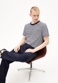 T-shirt a righe blu navy e bianco, con collo tondo e maniche corte. Pantaloni blu navy, seduto su una sedia marrone a trama con base in cromo.