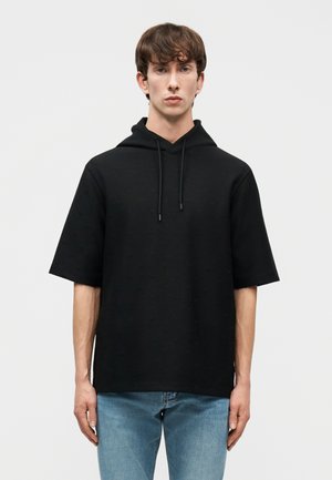 Mladý muž v čiernej mikine s krátkym rukávom a modrých džínsoch stojaci pred jednoduchým svetlosivým pozadím.