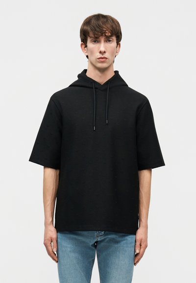 Jeune homme portant un hoodie noir à manches courtes et un jean bleu, debout devant un fond gris clair uni.