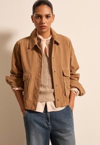 Beige Wildlederjacke mit Kragen, zwei Vorderaschen und hochgerollten Ärmeln, über einem strukturierten beigefarbenen Pullover und einem hellrosa Hemd getragen.