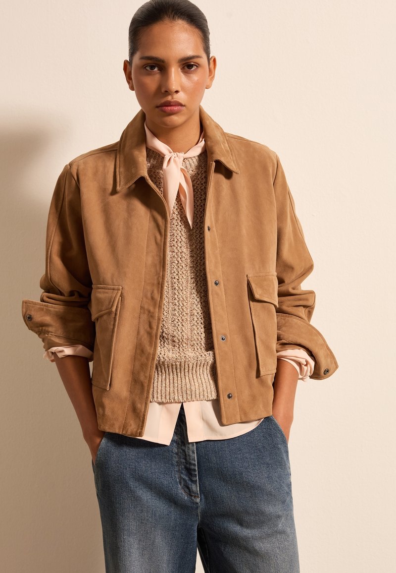 Beige Wildlederjacke mit Kragen, zwei Vorderaschen und hochgerollten Ärmeln, über einem strukturierten beigefarbenen Pullover und einem hellrosa Hemd getragen.