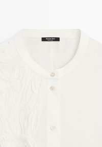 Chemise blanche à boutons avec col rond, motif texturé subtil sur un côté, et étiquette Massimo Dutti indiquant "Fabriqué en Inde".