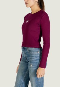 Vínový top s dlouhým rukávem a přiléhavým střihem, žebrovanou texturou a vyšívaným logem na hrudi, doplněný o džíny s vysokým pasem a potrhaným designem.