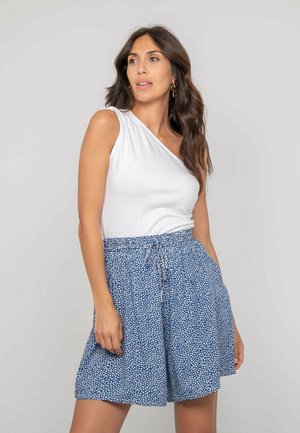 Femme portant un haut blanc à une épaule sans manches et un short taille haute bleu à motifs avec des poches, debout devant un fond uni.