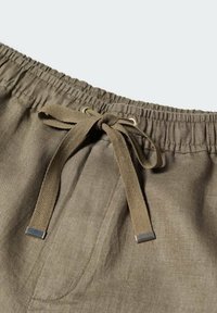 Ceinture de pantalon vert olive avec bande élastique et cordon de tissu noué avec embouts métalliques aux extrémités.