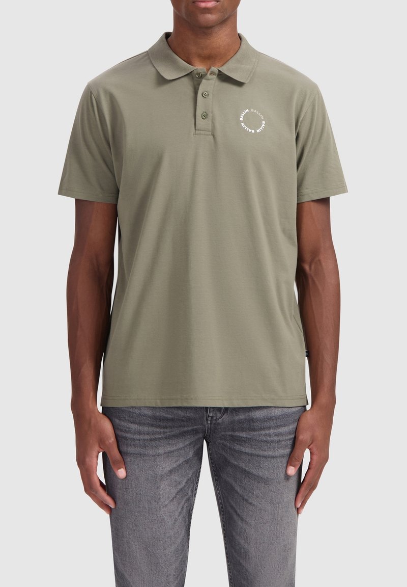 Khaki polo-shirt van katoen, met een klassieke kraag, drie knopen en een circulair logo op de linkerborst. Gekleed met grijze jeans.
