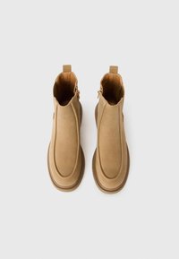 Pareja de botas de ante de color beige hasta el tobillo, con punteras redondeadas, cremalleras laterales y tiradores, fotografiadas desde arriba sobre un fondo blanco.
