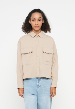 Beige kort skjorte med krage, to brystlommer foran og broderte detaljer, laget av et glatt stoff. Kombinert med lyse denimjeans.