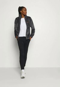 Pilkas vilnonis džemperis su aukštu apykakle, priekiniais užtrauktukais ir šoninėmis kišenėmis, derinamas su baltu marškinėliu ir juodomis kelnėmis; modelis dėvi baltus sportbačius.